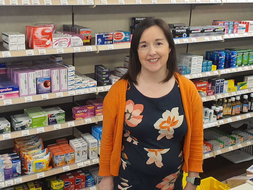 Photo of community pharmacist Emma Anderson smiling and standing in front of a display of medication