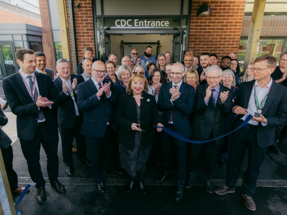 Group photo including representatives from Government, Sherwood Forest Hospitals Trust and other key partners. The group are standing outside the entrance to the Community Diagnostic Centre holding a ribbon and scissors.