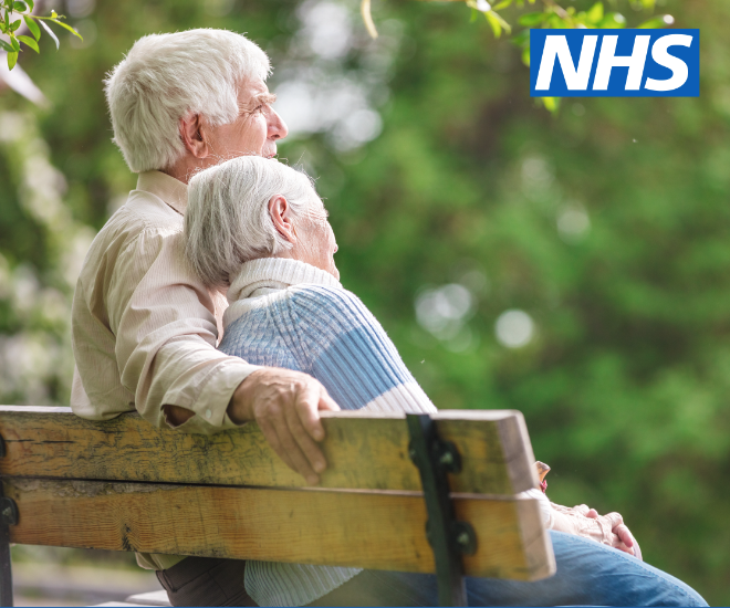 Two older adults sit on a park bench. The man has his arm around the woman.