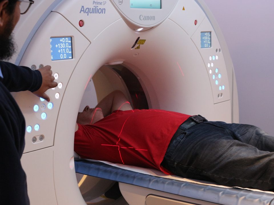 A man is lying down having a CT scan. A male radiographer presses a button on the scanner.