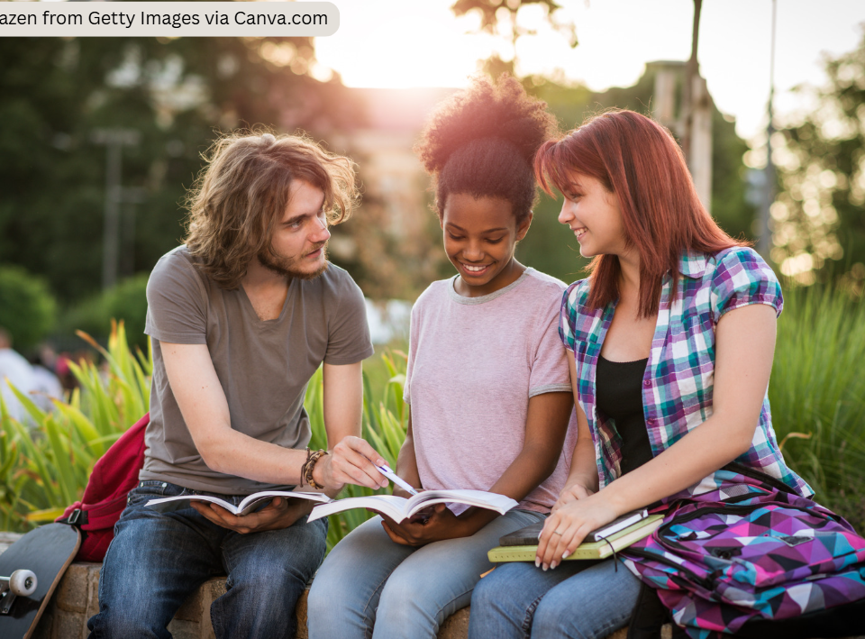 Three students sit outdoors on a low wall in a landscaped campus setting, studying together from open books and notebooks. One person points to a page while the others look on. Backpacks and a skateboard are beside them, with greenery and warm evening sunlight in the background.