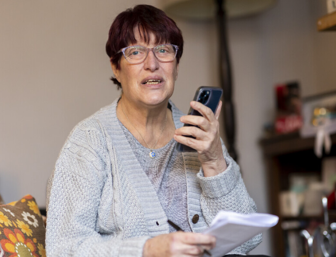 A person wearing glasses and a light grey knitted cardigan sits indoors holding a smartphone in one hand and a notepad with papers in the other. The person appears to be speaking into the phone while making notes. A patterned cushion is visible in the foreground, with a softly blurred living room setting in the background.